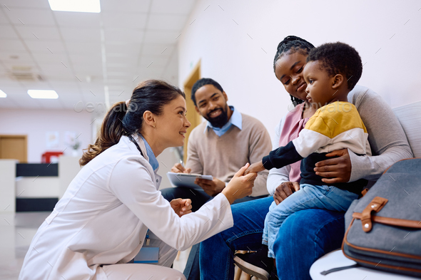 Female pediatrician talking to black little boy who is sitting on mother's lap at the clinic. - Stock Photo - Images