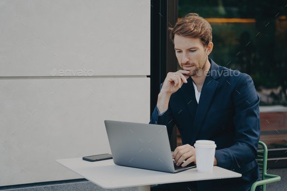 Handsome pensive young man browsing web on laptop while working remotely in sidewalk cafe - Stock Photo - Images