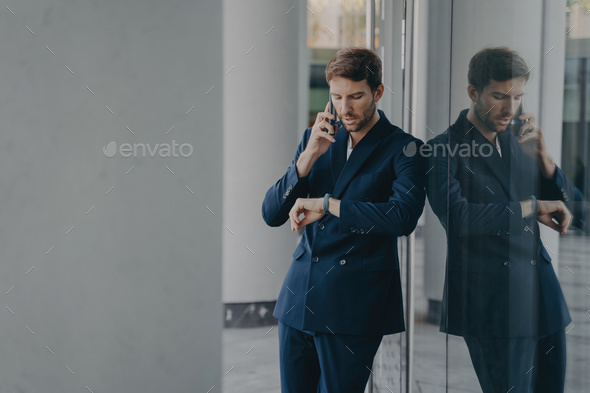 Pensive businessman in suit talking on mobile phone standing outside, looking at his hand watch - Stock Photo - Images