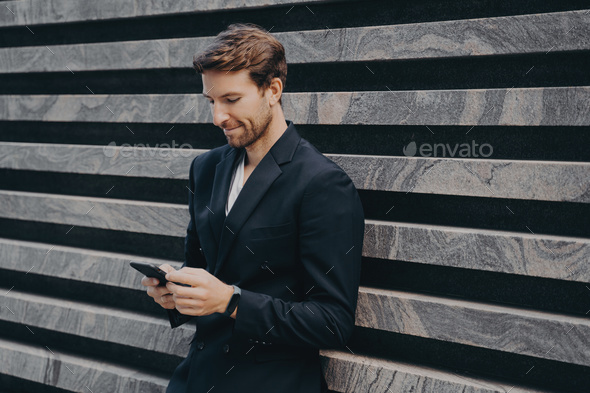 Pensive young office worker in formal stylish suit standing outdoor holding mobile phone in hands - Stock Photo - Images