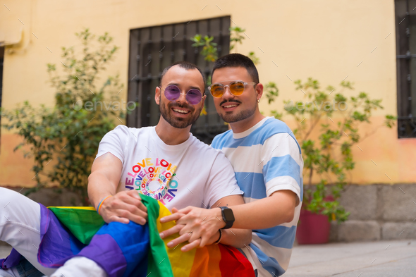 Portrait of gay male couple sitting on the floor smiling at pride party with rainbow flag, lgbt