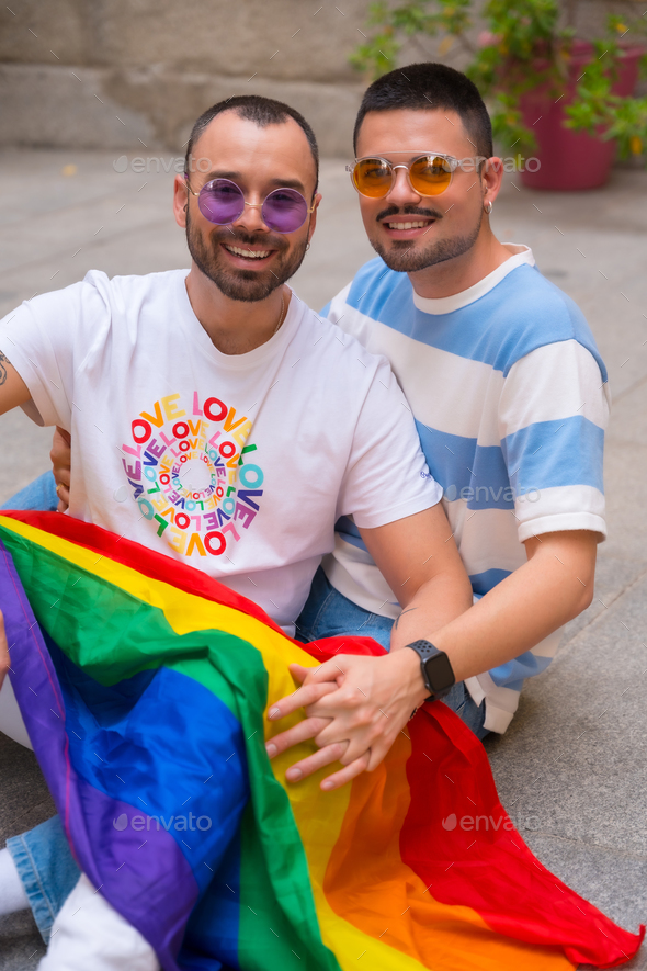 Portrait of gay male couple sitting on the floor kissing with rainbow flag at pride party, lgbt