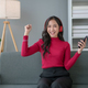 Happy young Asian woman raising hands with victory smiling happily with smart phone at home. - PhotoDune Item for Sale