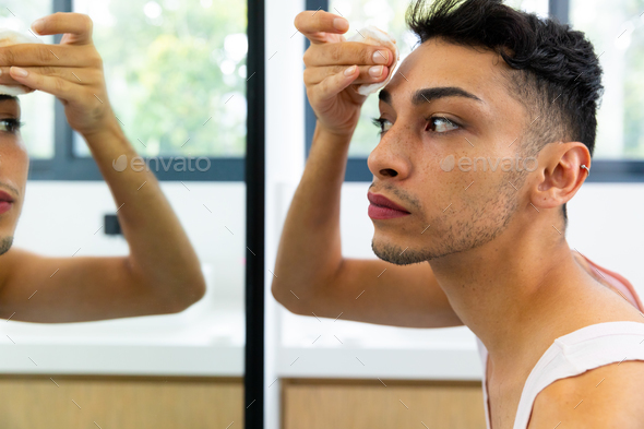 Biracial Transgender Man Looking In Mirror Using Cotton Pad For Cleansing Face In Bathroom 3001