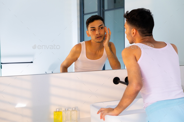 Biracial transgender man looking in mirror and touching face in ...