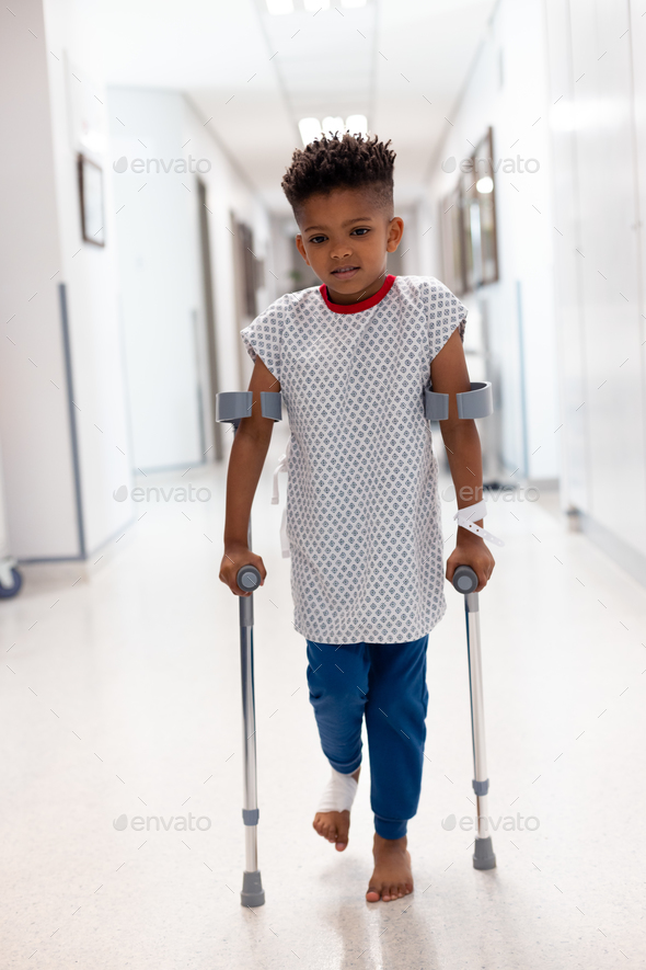 Vertical of african american boy patient walking with crutches in hospital corridor with copy space - Stock Photo - Images