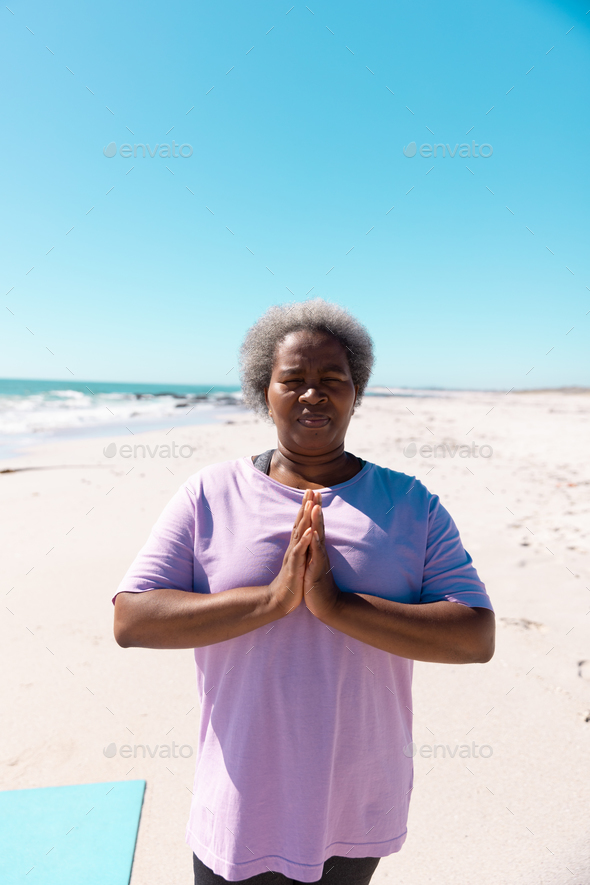 African american senior woman with short hair meditating in prayer pose at beach under clear sky