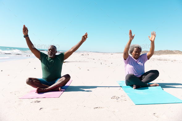 African american senior couple with arms raised meditating while sitting on mats at beach under sky