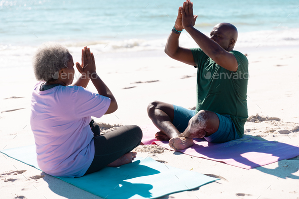 African american senior couple meditating in prayer pose on exercise mats at beach during summer