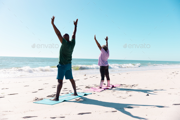 African american senior couple with arms raised exercising on mats at beach under clear blue sk