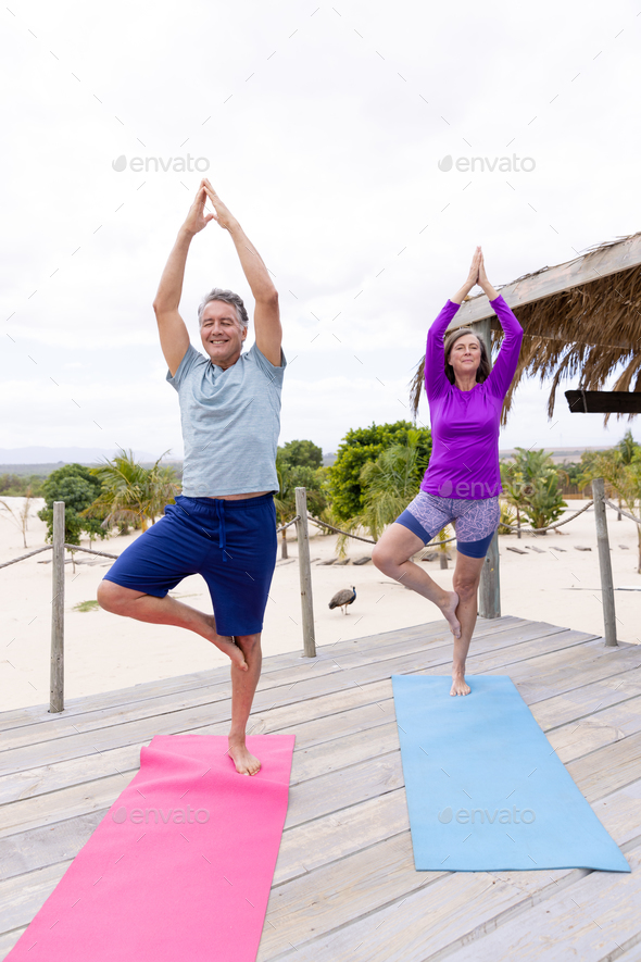 Caucasian senior couple with arms raised practicing tree pose on mats at beach in tourist resort