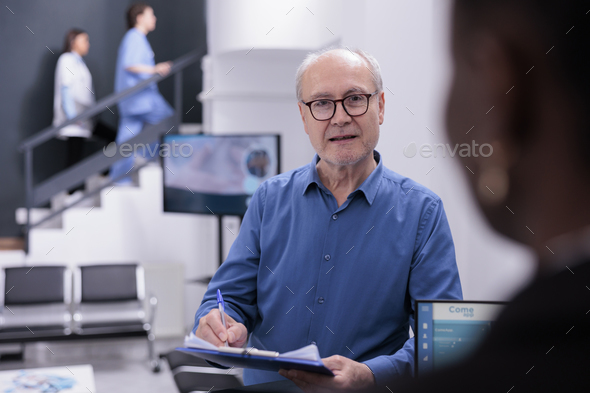 Caucasian man standing at reception counter asking receptionist for help with medical documents - Stock Photo - Images