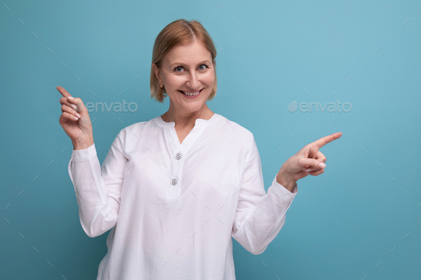 confident blond middle aged woman in white blouse pointing her finger to the side on studio