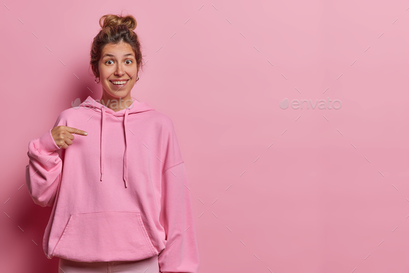 Photo of cheerful young woman dressed in comfortable sweatshirt points finger at blank space for