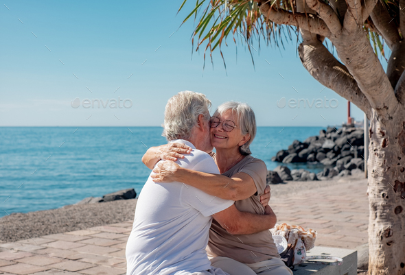 Lovely couple of senior people embracing with love sitting close to the sea. Elderly retirees joyful