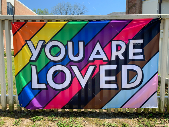 You are loved written on a progress pride flag integrated in one flag hanging on a fence