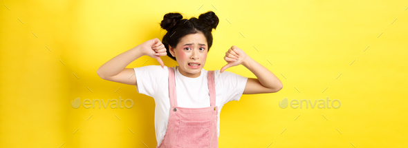 Disgusted asian girl showing thumbs down and cringe from bad product, standing in summer clothes