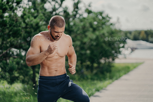 Active bearded man runs outdoors, listens to music, has a muscular body, poses near trees.