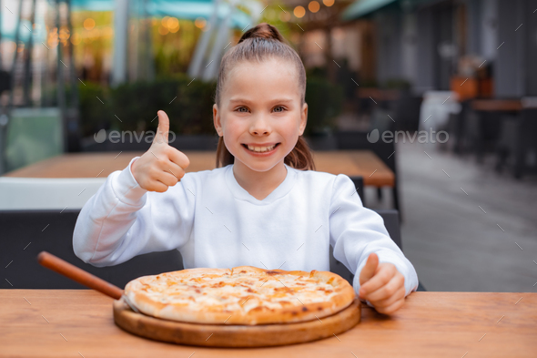 girl with hand good gesture and tasty pizza in pizzeria. child in white sweatshirt