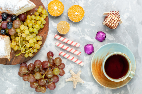 top view sugar powdered cake with fresh grapes candies and cup of tea on white desk fruit cake