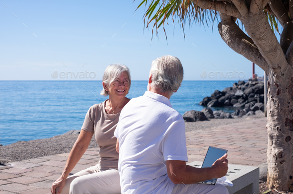 Happy couple of senior people sitting on bench close to the sea while man uses laptop.