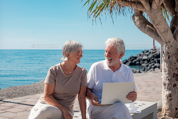 Cheerful senior couple sitting close to the sea in summer vacation. Elderly man using laptop.