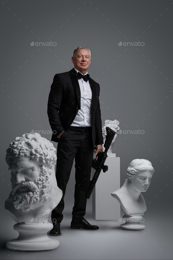 Stylish older man in expensive suit holding gun surrounded by ancient sculptures