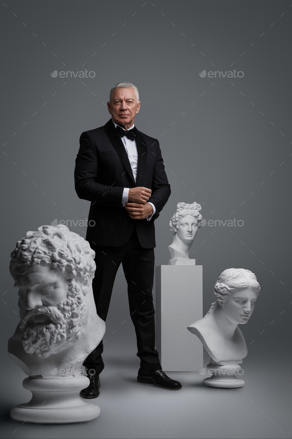 Stylish older man stands holding a gun, surrounded by three ancient sculptures
