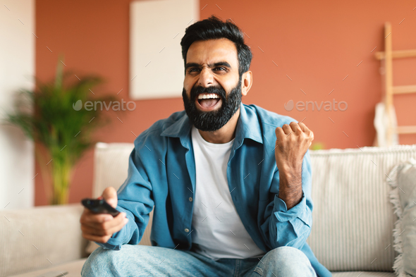 Joyful indian man shaking fists and shouting watching TV indoor