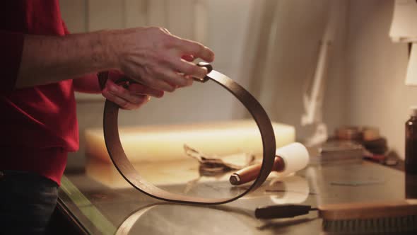A Man Ties Up the Brand New Leather Belt By the Table in a Workshop alt