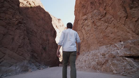 Low Angle Back View of Adult Man Walking on Road Through Narrow Gorge Between Tall Rocks alt