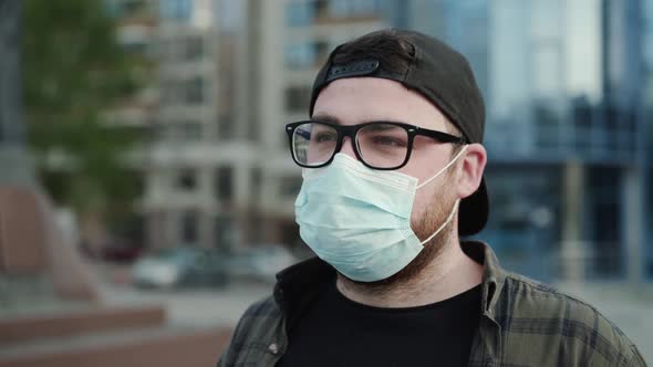 Portrait of Young Man in Glasses Cap and Medical Mask Blinks His Eyes Outdoors alt