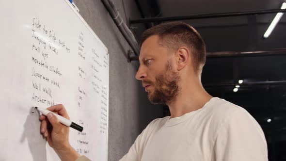 Athlete in White t Shirt Writes Down Exercises That He Will Perform on White Board with a Marker alt