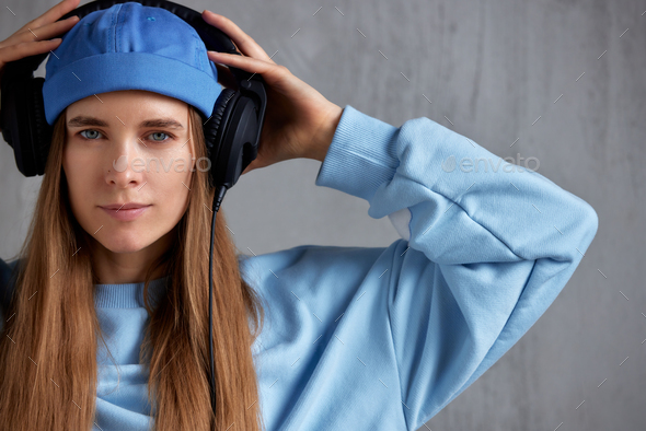 A young pretty long-haired girl in a blue sweater, a funny hat and black headphones listens to music