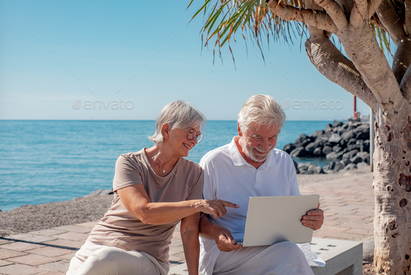 Cheerful senior couple sitting on a bench close to the sea using laptop together. Horizon over water