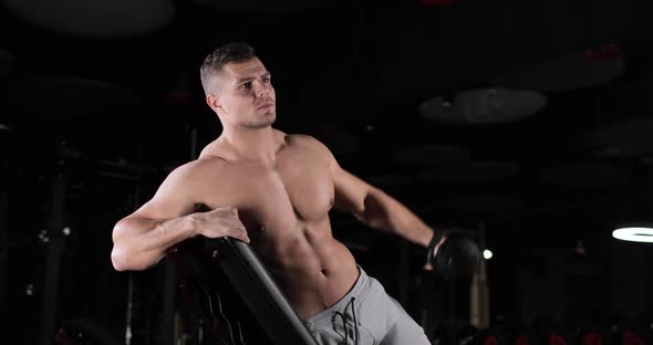 Young Athletic Man with a Naked Torso Doing Exercises with Dumbbells Leaning on a Bench in the Gym