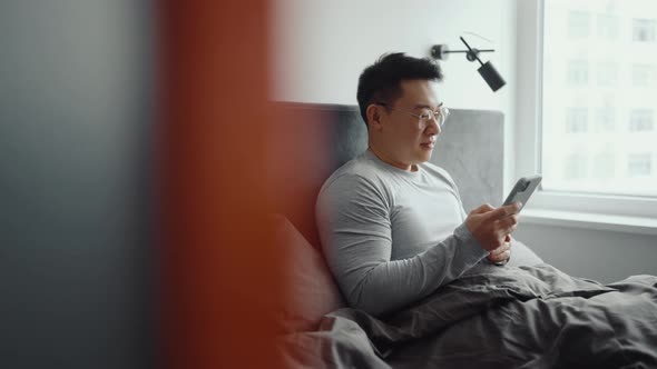 Happy Asian man wearing eyeglasses typing by mobile on the bed alt