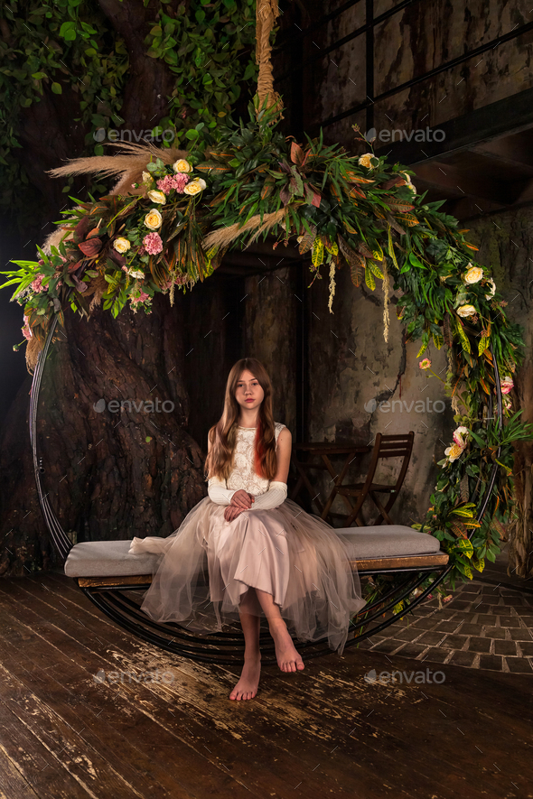 Teenager princess sitting on swing in light pink dress barefoot, posing in mystery room