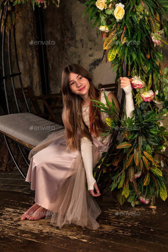 Teenager girl in light pink dress sitting on swing with flowers in mystery room