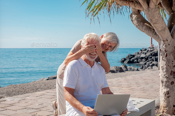 Playful senior couple sitting on a bench close to the sea using laptop together. Horizon over water