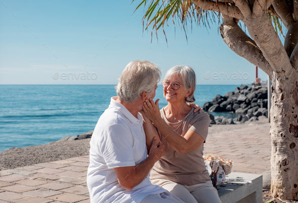 Happy bonding senior family couple sitting together on bench close to the sea cuddling