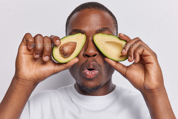 Surprised dark skinned man covers eyes with halves of avocado keeps mouth opened holds breath from