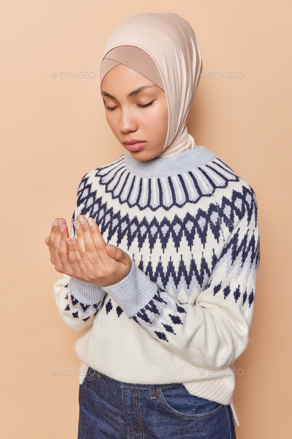 Calm young Muslim woman praying indoors wears headscarf and jumper standing with closed eyes making
