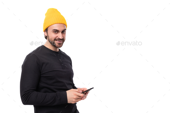 a shy young 30 year old guy with a beard dressed in a black jacket and a yellow hat is chatting on
