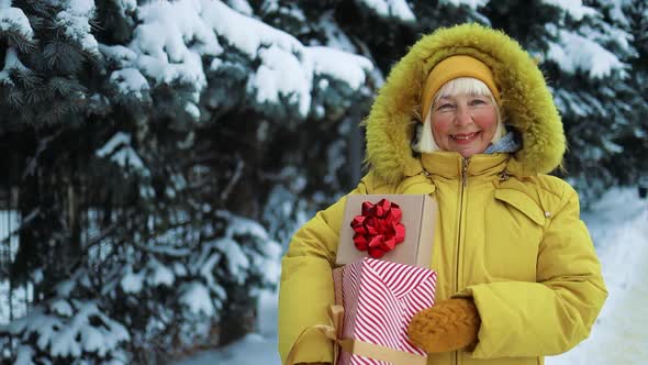 Positive Caucasian 50s Woman in Yellow Clothes Hands Holding Gift Boxes and Waving Hand on Christmas