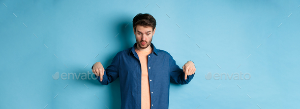 Image of shocked young man in casual clothes, looking and pointing down at something strange