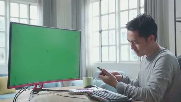 Asian Man With Green Screen Desktop Computer Using Mobile Phone While Working At Home alt