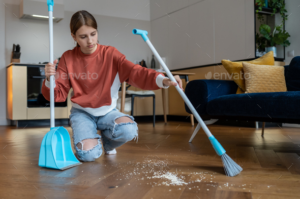 Focused teen girl sweep floor in living room from scattered debris with broom with brush and dustpan