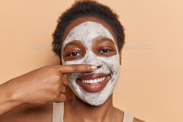 African curly haired woman wearing facial cosmetic mask exudes cheerfulness as she joyfully smiles