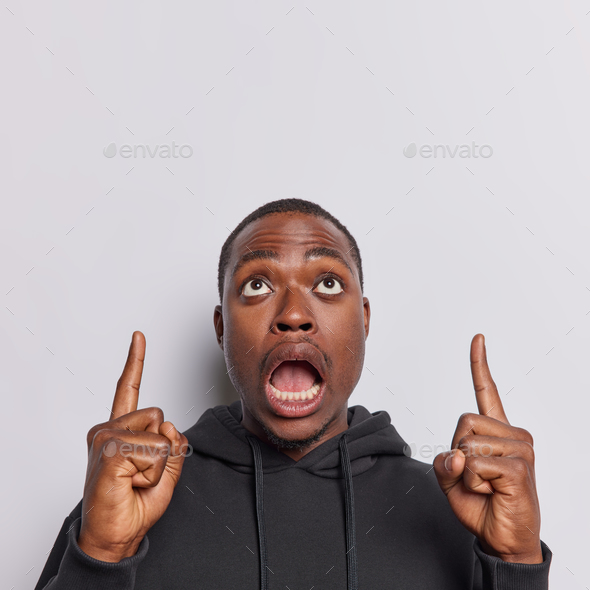 Stupefied African man dressed in casual attire points upwards with wide eyed shocked expression his